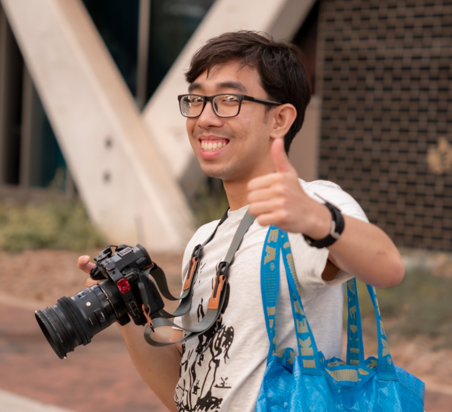 Poom holding camera, giving thumbs up, wearing bag.